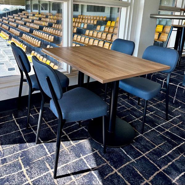 A modern rectangular dining table with a light wood top and black metal pedestal base, surrounded by four blue upholstered chairs with black frames, set on a dark patterned carpet in a bright indoor space with large windows.